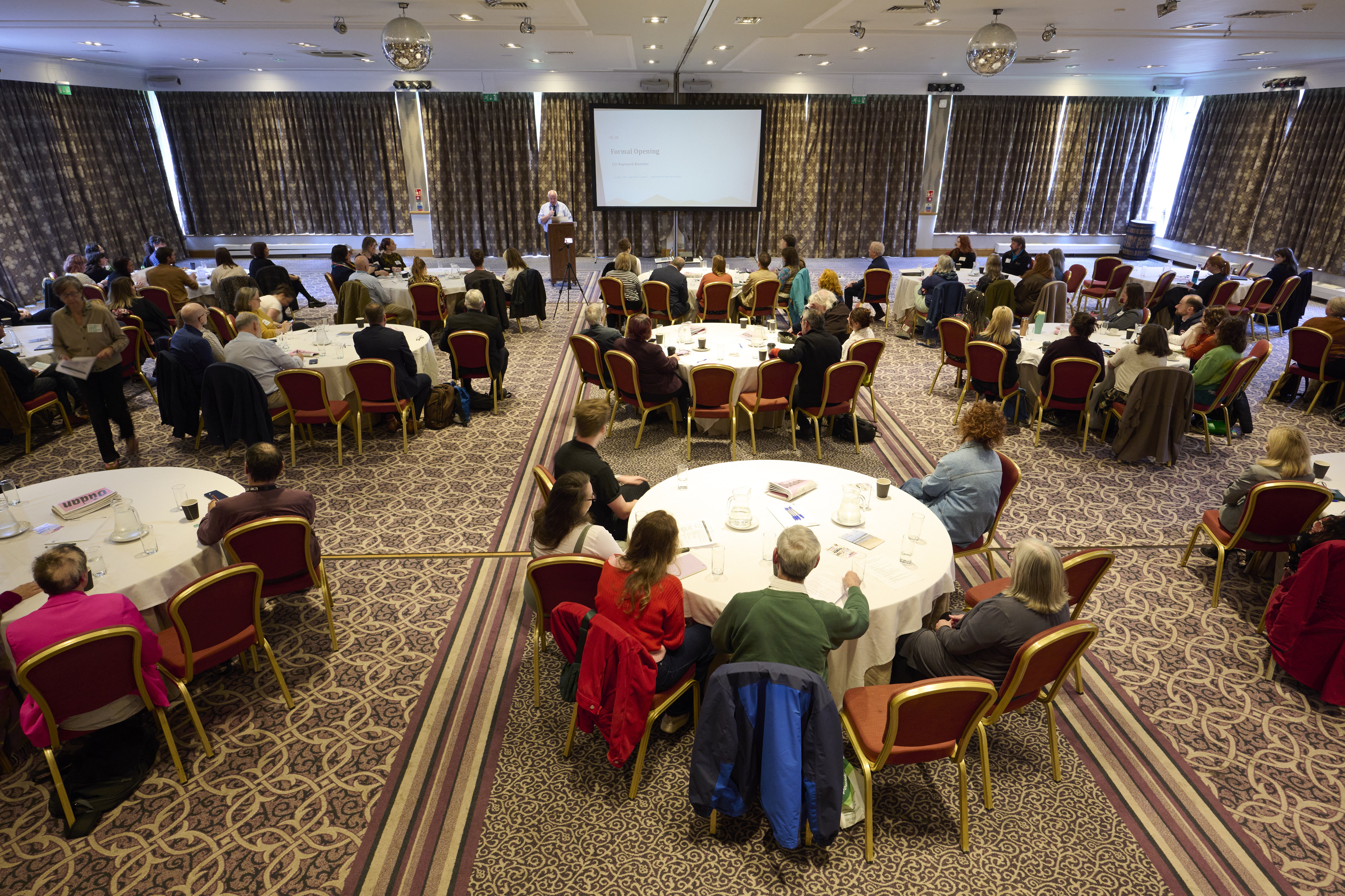 More than 100 people attend the Highlands’ first Cultural Assembly. The image shows people gathered around tables in a conference venue and listening to a speaker leading discussions on the future of culture in the region.